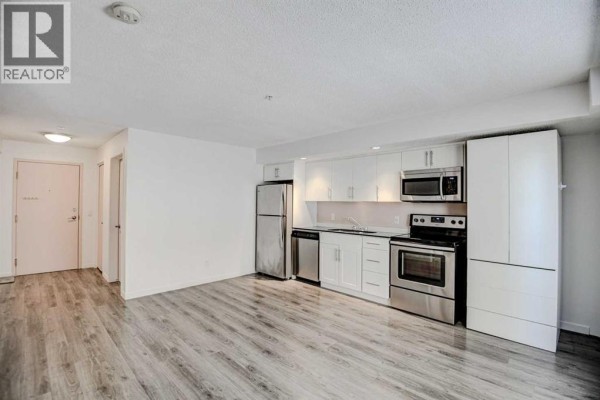 View of an open-concept living area with a kitchen, white cabinets, and grey wood-look flooring. - 106, 1920 11 Avenue Sw, Calgary