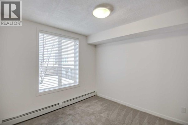 Bedroom with carpeted floor, large window with blinds, and white walls. - 106, 1920 11 Avenue Sw, Calgary