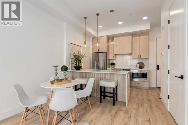 Open-concept dining area with a small round table, four white chairs, and view into the kitchen. - 106, 330 Dieppe Drive Sw, Calgary