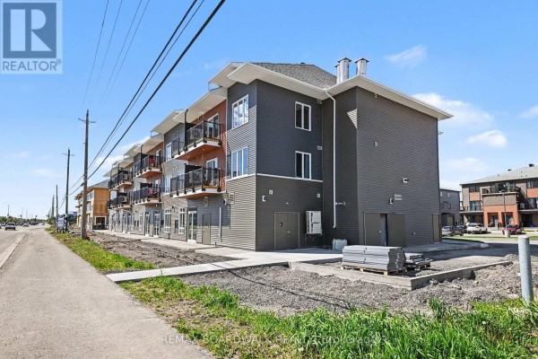 Rear exterior view of a three-story commercial building with gray siding, storage doors, and a sidewalk. - 106 - 2370 Tenth Line Road, Ottawa