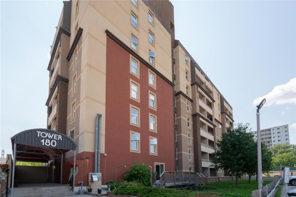 Exterior view of Tower 180 apartment building with a covered entrance and surrounding greenery. - 106 180 Beliveau Road, Winnipeg