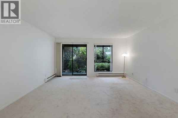 View of an empty living room with beige carpeting and patio doors opening to a lush garden. - 106 333 Wethersfield Drive, Vancouver