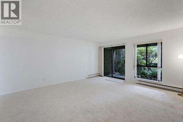 Spacious living area with beige carpet, patio doors, and a window showcasing abundant green foliage. - 106 333 Wethersfield Drive, Vancouver