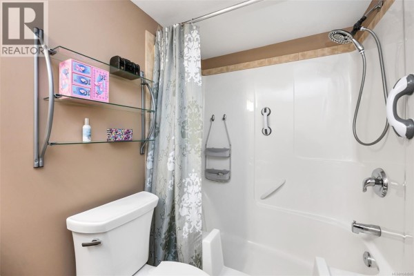 Bathroom with a white bathtub, shower curtain, toilet, and glass shelves. - 106 650 Dobson Rd, Duncan