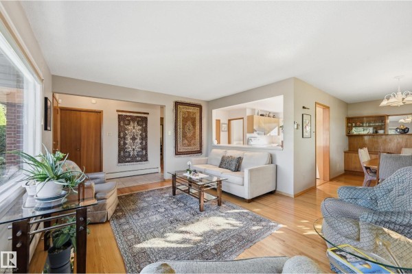 Living room with hardwood floors, beige walls, two sofas, multiple armchairs, and a pass-through to the kitchen. - 10607 74 St Nw, Edmonton