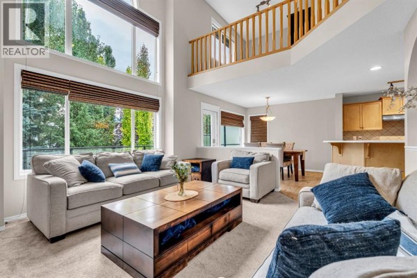Living room with a high ceiling, large windows, neutral furniture, and a view of trees. - 1062 Tuscany Drive Nw, Calgary