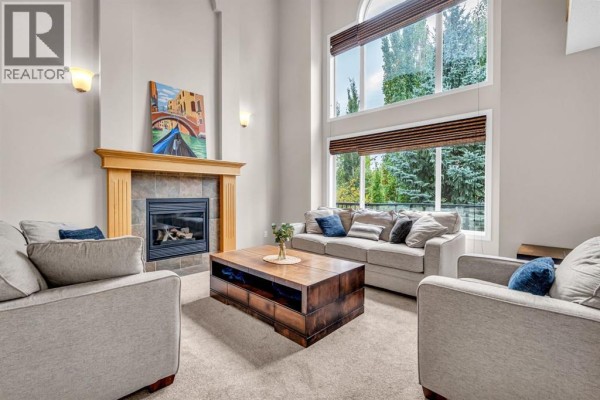 Living room with a fireplace, wood mantle, tile surround, and large windows with tree views. - 1062 Tuscany Drive Nw, Calgary