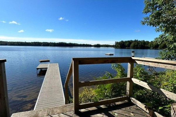 View from a wooden deck overlooking a lake with two docks and a clear blue sky. - 1063 Dickie Lake Road, Lake Of Bays