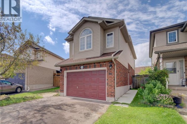 Side exterior view of a two-story house with tan siding, brick base, and a red garage door. - 1064 Copper Leaf Crescent, Kitchener