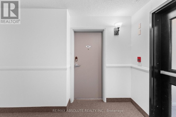 Apartment hallway with a beige door labeled 107 and brown carpeted floor. - 107 - 16 Sheffield Street, Arnprior