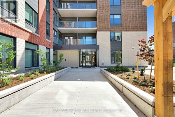 Glass entrance to the apartment building with concrete walkways and landscaping. - 107 - 385 Hill Street, London East