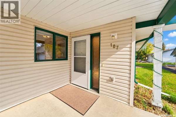 Entrance door of unit 107 with a beige doormat on a concrete porch. - 107 111 2nd Avenue E, Rosetown