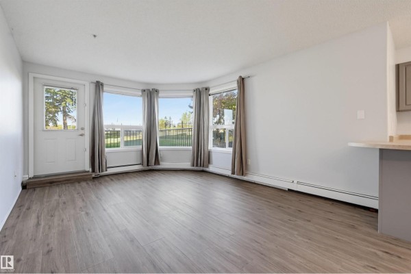Living area with a bay window, grey curtains, and a white door leading outside. - #107 13635 34 St Nw, Edmonton