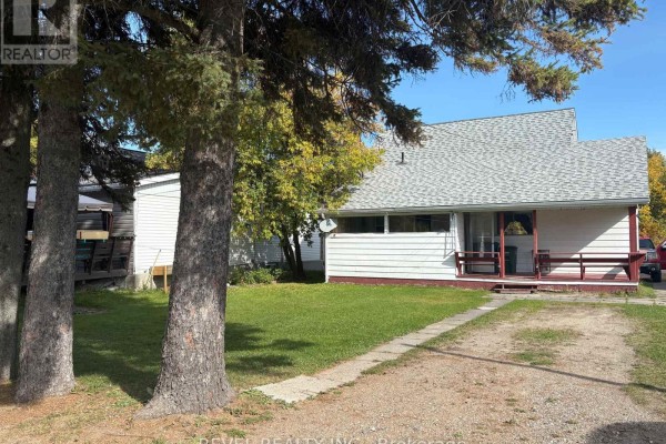 View of the house from the side yard, framed by large pine trees and a gravel driveway. - 107 Fifteenth Avenue, Cochrane