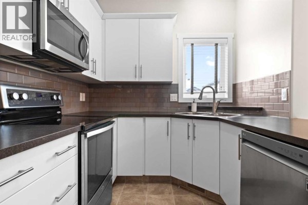 Corner view of the kitchen showing dark countertops, white cabinets, stainless steel dishwasher, and a window above the sink. - 107 Henderson Crescent, Penhold