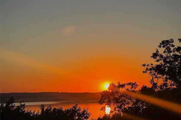 Sunset over a calm lake, with silhouetted trees in the foreground and orange light rays. - 107 Sunset Drive, Lake Of The Prairies