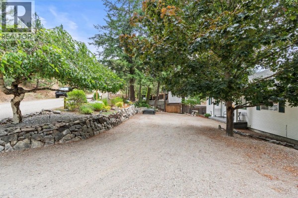 Gravel driveway alongside a stone wall and trees, with the side of a white house visible. - 10771 Pinecrest Road, Vernon