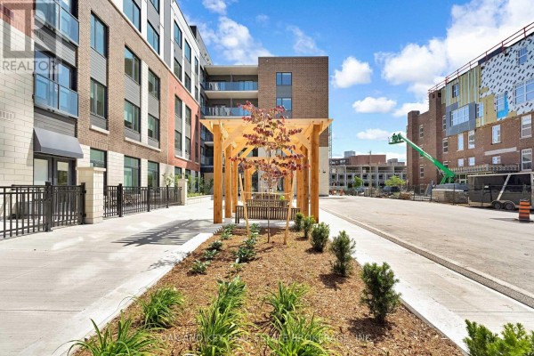 Side view of the apartment building entrance with a paved walkway, wooden pergola, and surrounding landscaping. - 108 - 385 Hill Street, London East