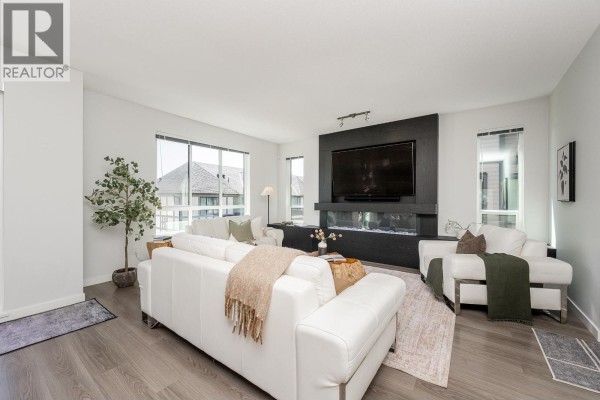 Living room with white leather sofas, a dark wood feature wall with a television, and large windows. - 108 3500 Burke Village Promenade, Coquitlam