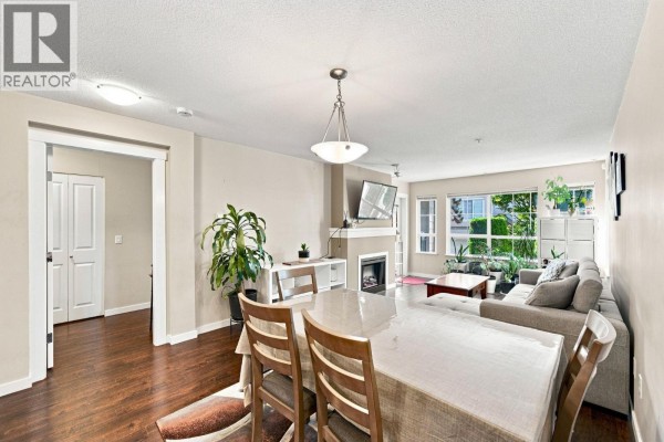 Living and dining area with a fireplace, sofa, coffee table, dining table with chairs, and wooden flooring. - 108 9233 Government Street, Burnaby