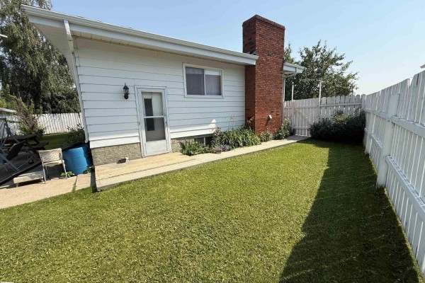 View of the backyard with a green lawn, white fence, and exterior brick chimney. - 10808 109 Street, Westlock