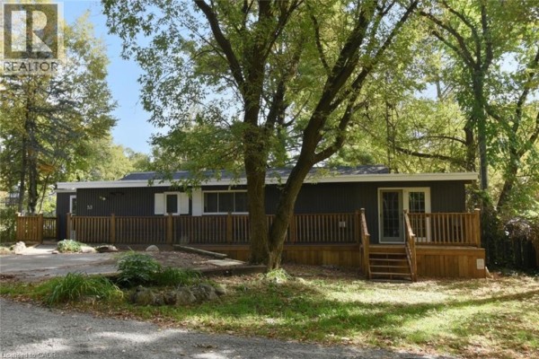 Front view of the dark-sided mobile home with a wrap-around wooden deck and trees. - 1085 W Concession 10 Concession W Unit# 99, Flamborough