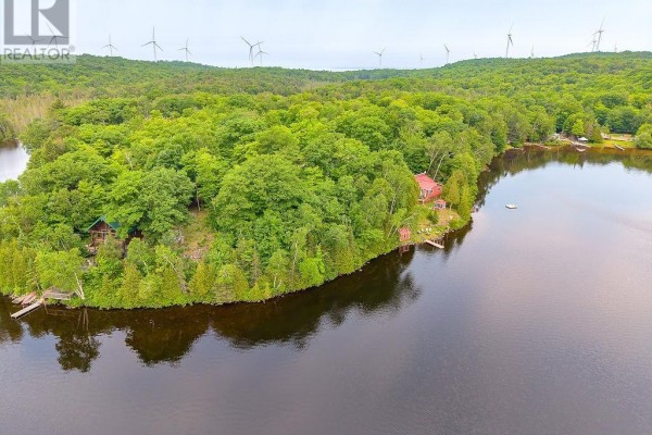 Aerial view of a tree-covered island with multiple buildings, docks, and windmills on distant hills. - 1089 Prince Lake Rd, Prince Township