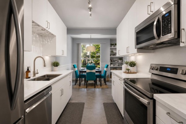 Galley kitchen with stainless steel appliances, white countertops, and a dining area with teal chairs visible. - 109 14980 101a Avenue, Surrey