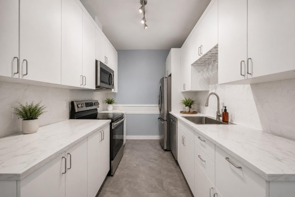 Long galley kitchen with white cabinets, stainless steel appliances, and marble-patterned countertops. - 109 14980 101a Avenue, Surrey