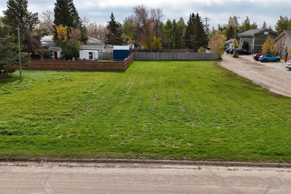 A wide, grassy vacant lot with a wooden fence in the background, adjacent to a paved street and houses. - 109 1st Street, Swan River