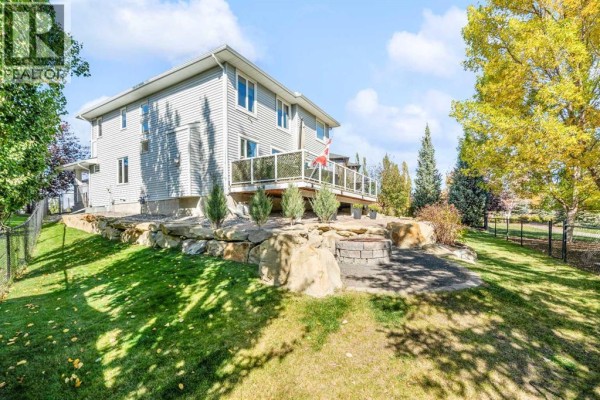 Rear view of a two-story house with a wooden deck and a stone fire pit area. - 109 Drake Landing Cove, Okotoks