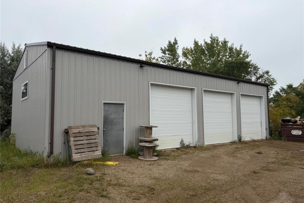 Rear exterior of a grey metal building with three white garage doors, a metal door, and wooden pallets. - 109 Industrial Drive, Brandon
