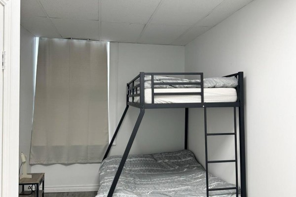 Bedroom with grey patterned bedding on black metal bunk beds, a window with light grey curtains, and a bedside table. - 11 King Street E, Cobourg