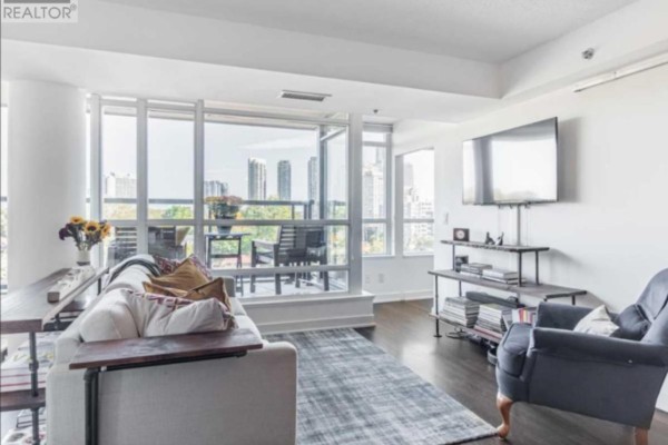 Living room with a large sofa, armchair, television, and floor-to-ceiling windows overlooking a balcony and cityscape. - 11 Superior Ave Avenue, Toronto