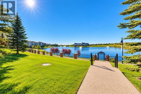 Walkway leading to a lake with fountains and apartment buildings in the distance. - 110, 162 Country Village Circle Ne, Calgary