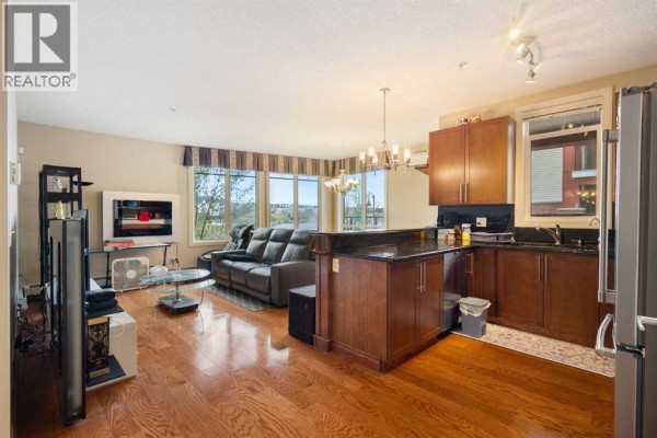 Living room and kitchen area with dark leather sofa, island, and hardwood floors. - 110, 162 Country Village Circle Ne, Calgary