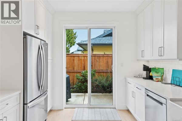 View through a sliding glass door from a white kitchen to a fenced backyard with greenery. - 110 3003 Keystone Dr, Duncan