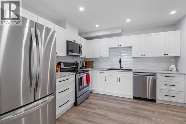 Modern white kitchen with stainless steel appliances, grey countertops, and wood-look flooring. - 110 455 Killoren Crescent, Prince George