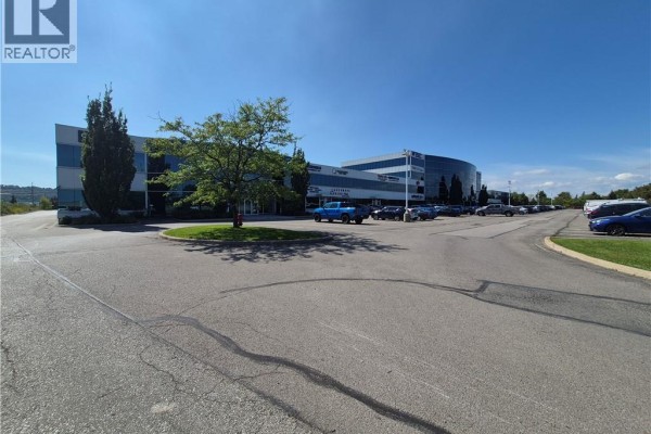Exterior view of a large commercial building with a circular driveway, trees, and a parking lot. - 1100 South Service Road Unit# 114, Stoney Creek