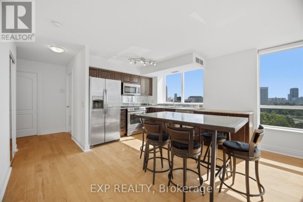 Kitchen with dark cabinets, stainless steel appliances, island with bar stools, and adjacent living area with TV. - 1103 - 1103 Leslie Street, Toronto