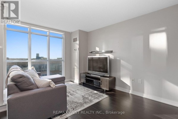 Living room with grey sectional sofa, television, and large window overlooking the city. - 1106 - 15 Iceboat Terrace, Toronto