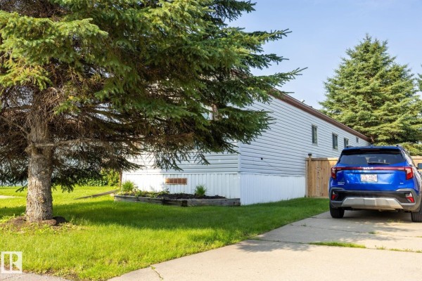 View of a mobile home with white siding, a blue SUV, and a large evergreen tree. - 1106 Lake Vista Dr, Sherwood Park