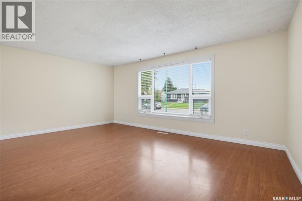 Living room with wood laminate flooring, cream walls, and a large window facing the street. - 1108 Tiffin Crescent, Saskatoon