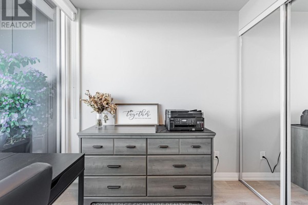 Close-up of a grey dresser with decorative items and a printer, next to a mirrored closet door. - 1114 - 7895 Jane Street, Vaughan