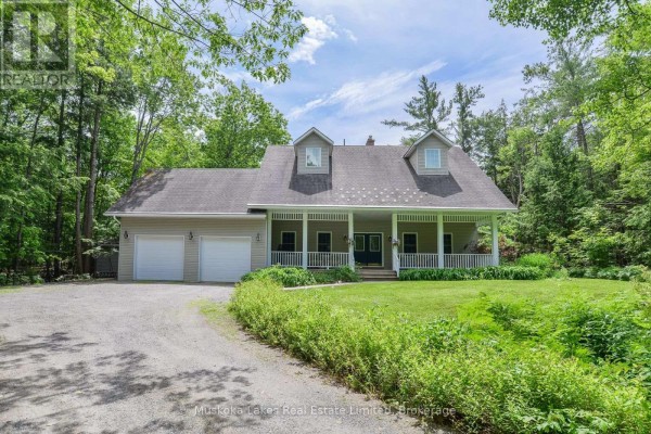 Front exterior of a tan vinyl-sided house with a wraparound porch, two-car garage, and surrounded by trees. - 1115 Partridge Lane, Bracebridge)