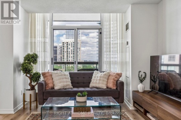 Gray sofa with pink and white pillows in front of a large window with city view. - 1116 - 503 Beecroft Road, Toronto