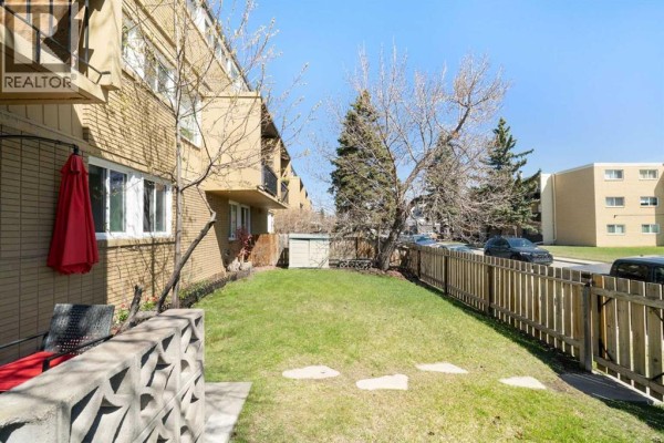 Private grassy backyard area with a wooden fence, trees, and a patio. - 112, 7007 4a Street Sw, Calgary
