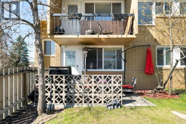Ground floor patio with a barbecue, chairs, and a red umbrella in front of an apartment unit. - 112, 7007 4a Street Sw, Calgary