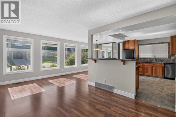 Living room with large windows overlooking the yard, wood flooring, and open pass-through to kitchen. - 112 Beaconsfield Rise Nw, Calgary