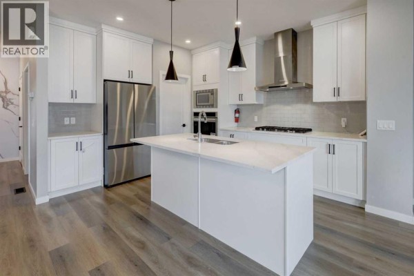 Modern white kitchen with a large island, stainless steel appliances, and light grey wood-look flooring. - 112 Emberside Place, Cochrane